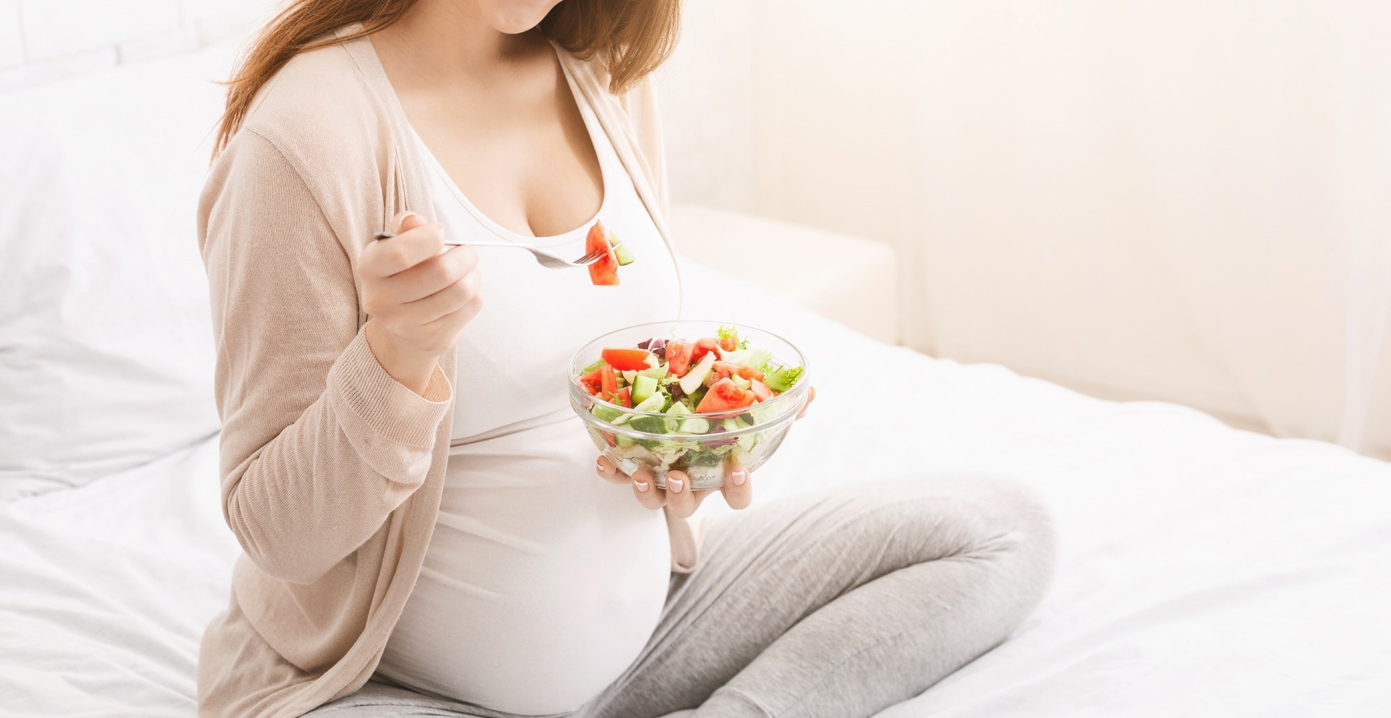A pregnant woman is sitting on a bed, holding a bowl of salad in her hands