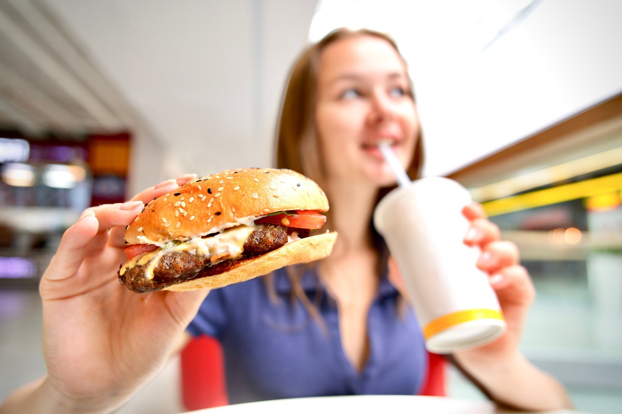 young woman holding big burger and cup of soda in shopping mall on food court.