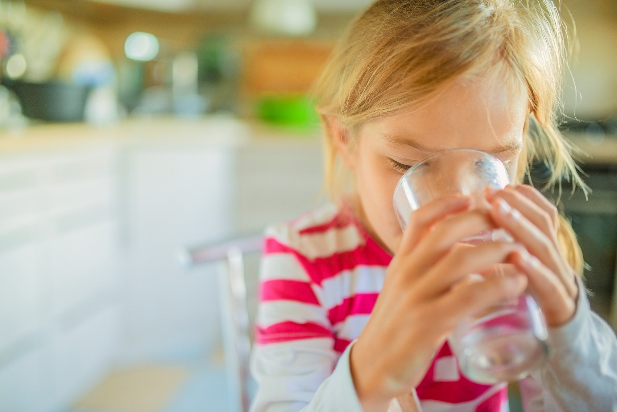 Beautiful smiling little girl drinking a glass of water against the background of the kitchen.