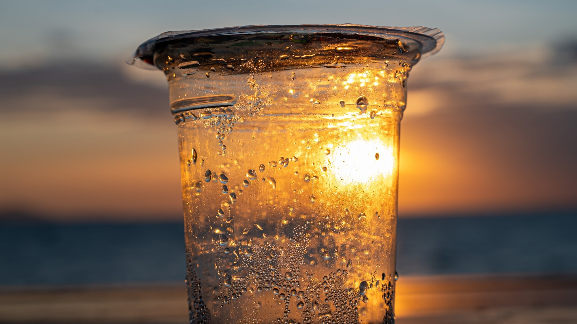 A plastic cup with water droplets reflects the golden sunset over the ocean